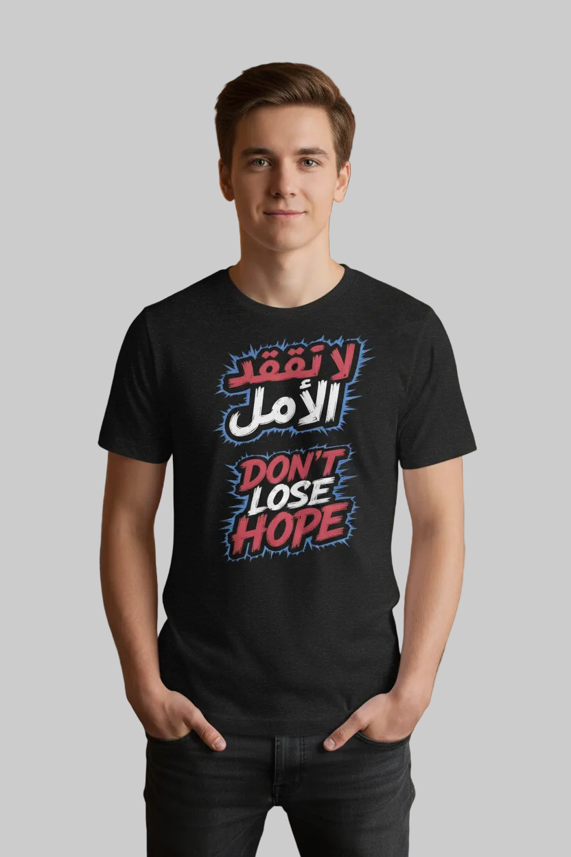 A young man stands in a cafe wearing a black t-shirt with a bold graphic print in Arabic and English that reads 'Don't Lose Hope' in red, white, and blue. The background shows a warm, softly blurred coffee shop interior with wooden tables, hanging light bulbs, and shelves.